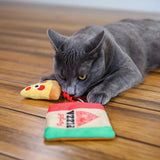 A gray cat lies on the wooden floor, engrossed in playing with Mad Cats Purrfect Pizza, a plush puzzle toy shaped like a pizza slice and box. Infused with North American catnip, the toy captivates the cat as it paws curiously.