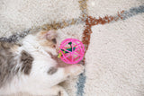 A fluffy white and gray cat lies on a textured rug, playfully batting the Quirky Kitty Guacmeowy Ball of Fury, a pink toy with a green feather that mimics an electronic cat toy.