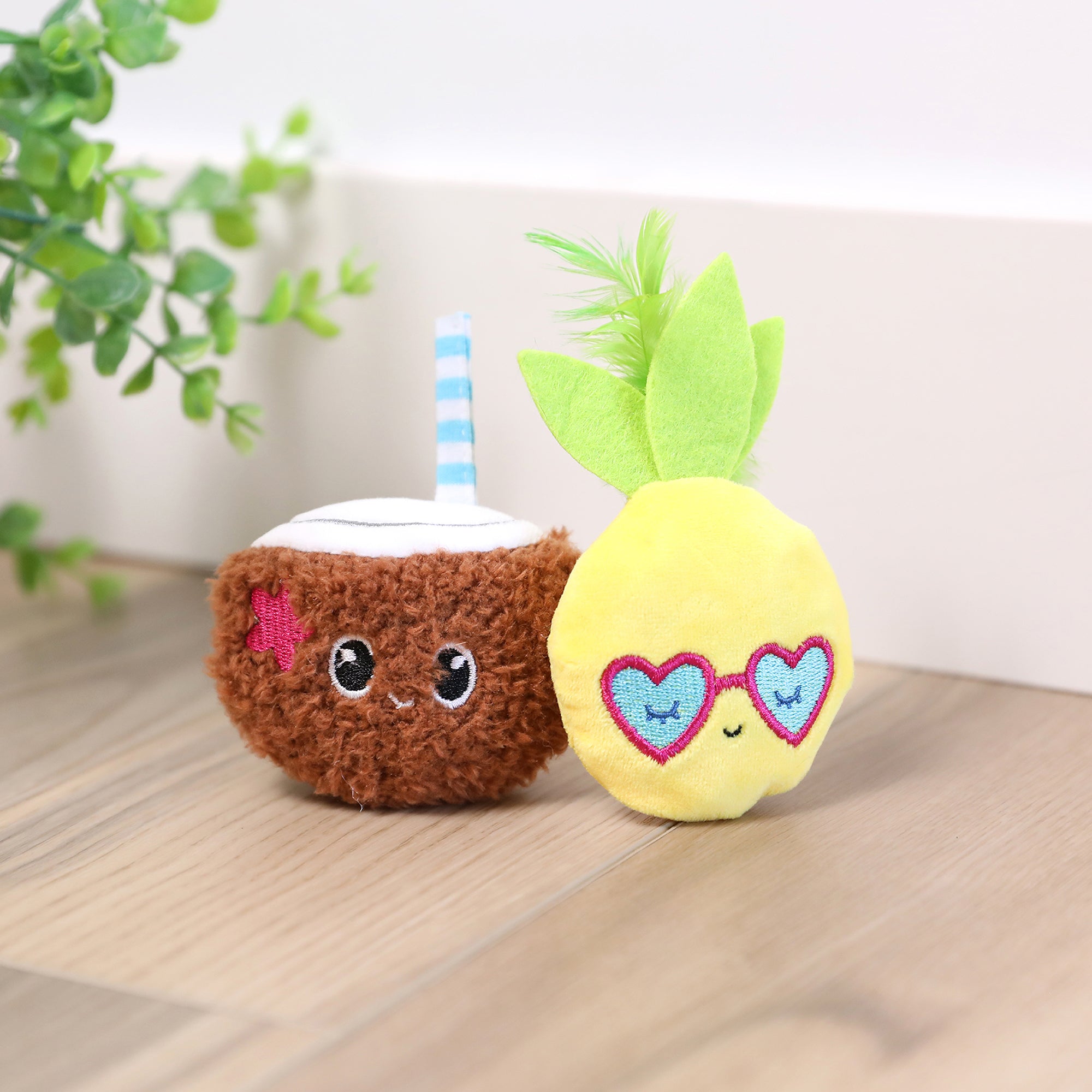 On a wooden floor sit two plush toys: a smiling brown coconut adorned with a pink flower and blue straw, and a yellow pineapple with heart-shaped sunglasses. Partially visible on the left is the Quirky Kitty Purrfect Pina Colada Catnip Plush Cat Toy, adding charm to the playful scene.