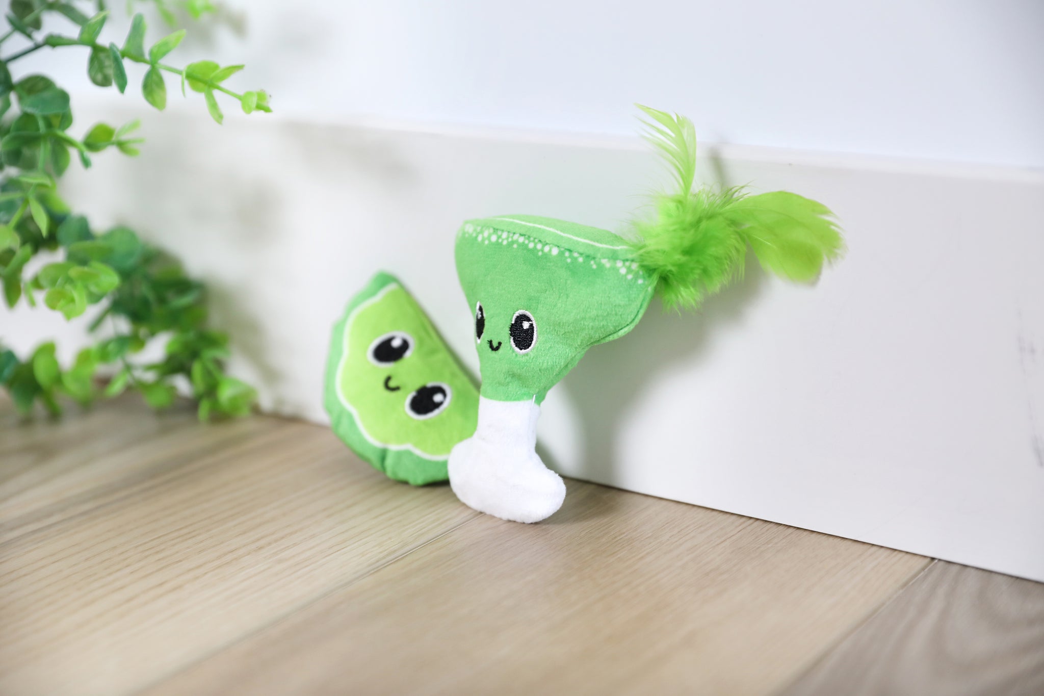 Two adorable, green Quirky Kitty Meowgarita Time Catnip Plush Toys, resembling fruits or veggies with smiling faces and googly eyes, rest on a wooden floor. One features a feather on top. Greenery decorates the left side of the image, complemented by playful hints of crinkle paper.