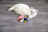 A fluffy white cat with blue eyes playfully holds the Mad Cat Canned Ham, a blue and yellow cat-shaped toy sprinkled with silvervine, lying on a wooden floor beside a charming pink pig toy.