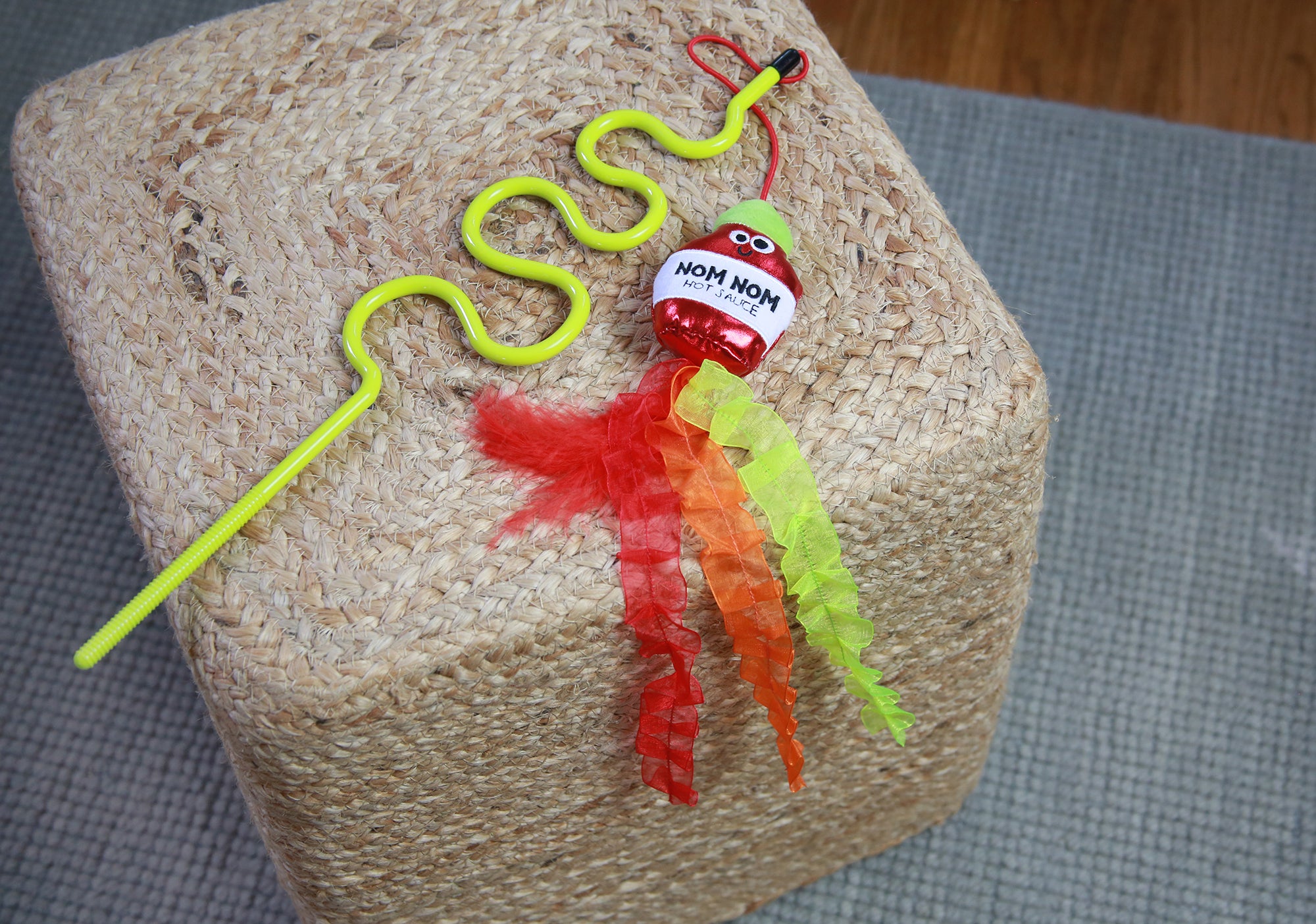 The Mad Cat® Nom Nom Hot Sauce Ribbon Wand, a yellow wavy plastic stick toy with Nom Nom in red, lies on a woven square ottoman. It has bright red, orange, and green crinkly strips and sits on a blue-gray rug.