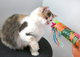 A fluffy white and gray cat sits on a dark surface, intently watching the Quirky Kitty® Peek & Pounce Wand by Quirky Kitty, adorned with colorful ribbons and feathers, being held toward it.
