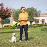A young person in a yellow t-shirt stands on the grass, holding a Hyper Pet K9K2 Kannon Mini Tennis Ball Launcher. A small beagle eagerly sits beside them. In the background, theres a building and trees with autumn leaves.