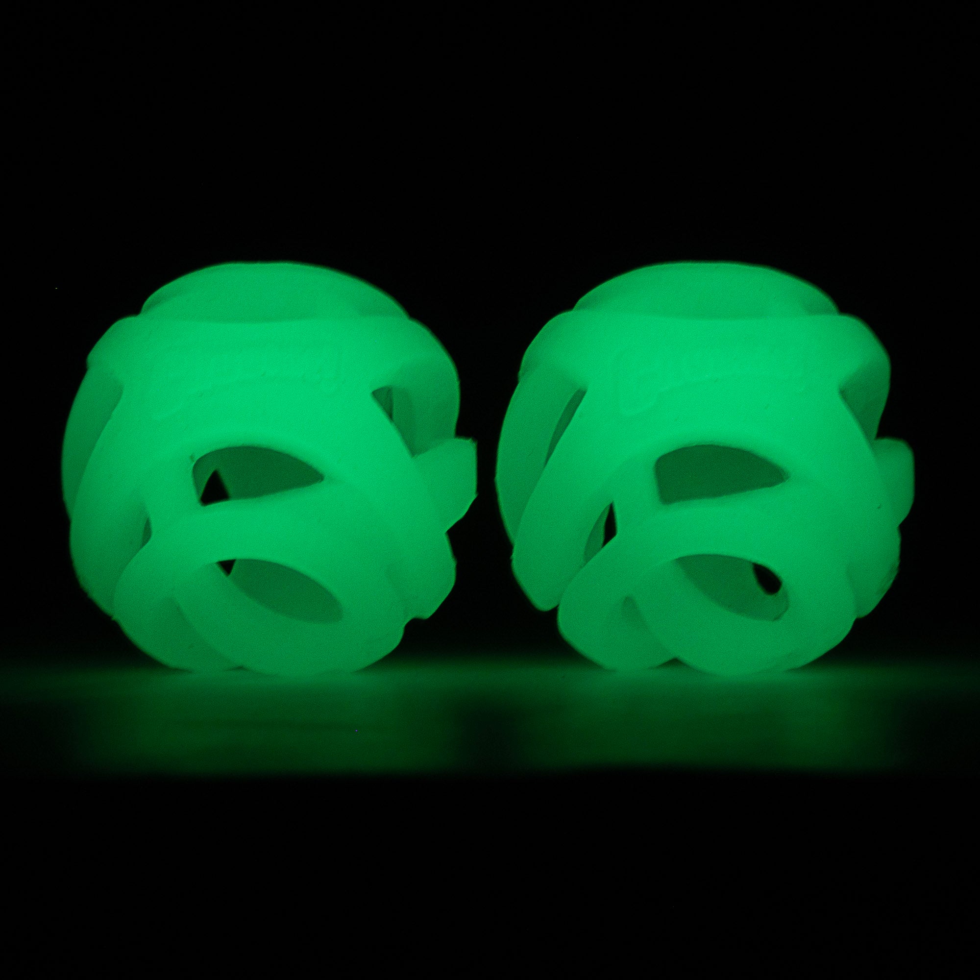 Two round, green Chuckit! Max Glow Air Fetch Balls with interwoven patterns are shown against a black background, emitting a soft luminescent light for dog fetch fun.
