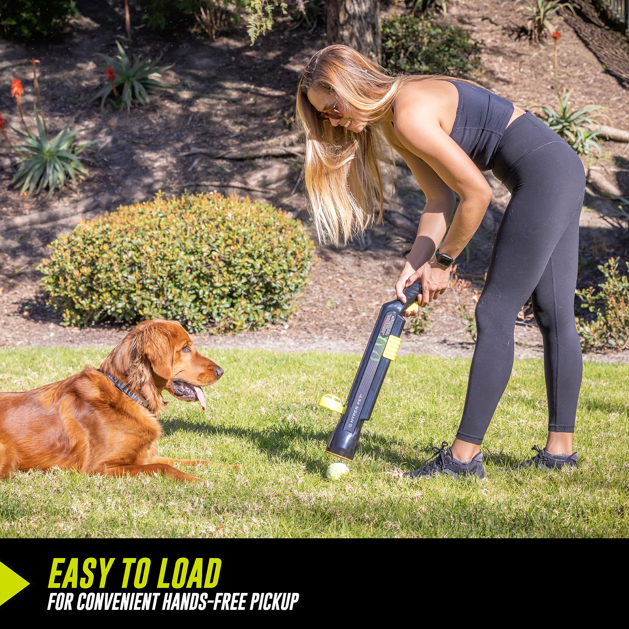 A woman in athletic wear uses the Hyperpet K9 Kannon Pro Ball Launcher to easily pick up a tennis ball while her brown dog watches on a grassy lawn. Text overlay reads, Easy to load for convenient hands-free pickup.
