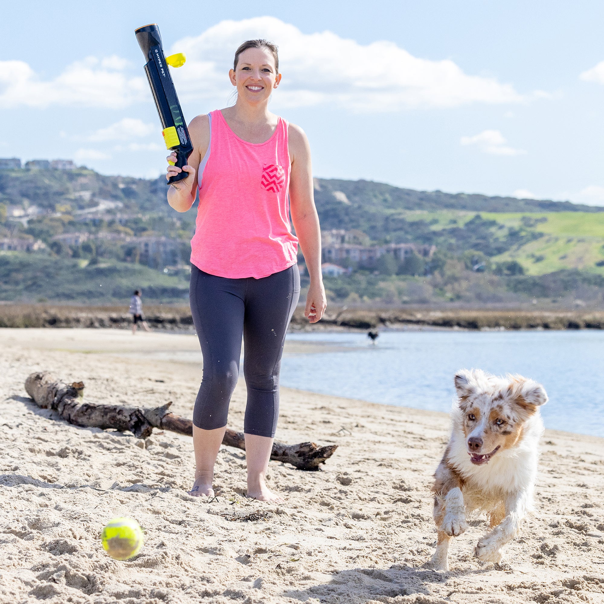 Wearing a pink tank top and gray leggings, a woman enjoys beach playtime with her dog using the Hyperpet K9 Kannon Pro Ball Launcher. She launches a yellow tennis ball, prompting her eager pup to chase it across the sand, all against rolling hills and a bright blue sky.