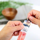 Hands are opening a packet of Wild Eats Simply Puree Ahi Tuna Cat Treat, featuring coral packaging with white and black text. It displays an image of Ahi Tuna and promises natural ingredients. The sustainably sourced Omega-3 rich fish sits against a wooden bowl backdrop with greenery.