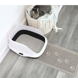 A gray tabby cat sits in a round basket with a striped blanket next to a white litter box filled with cat litter. The Arm & Hammer Paws Litter Mat is on the gray wooden floor, preventing scatter and showcasing its durable rubber material.