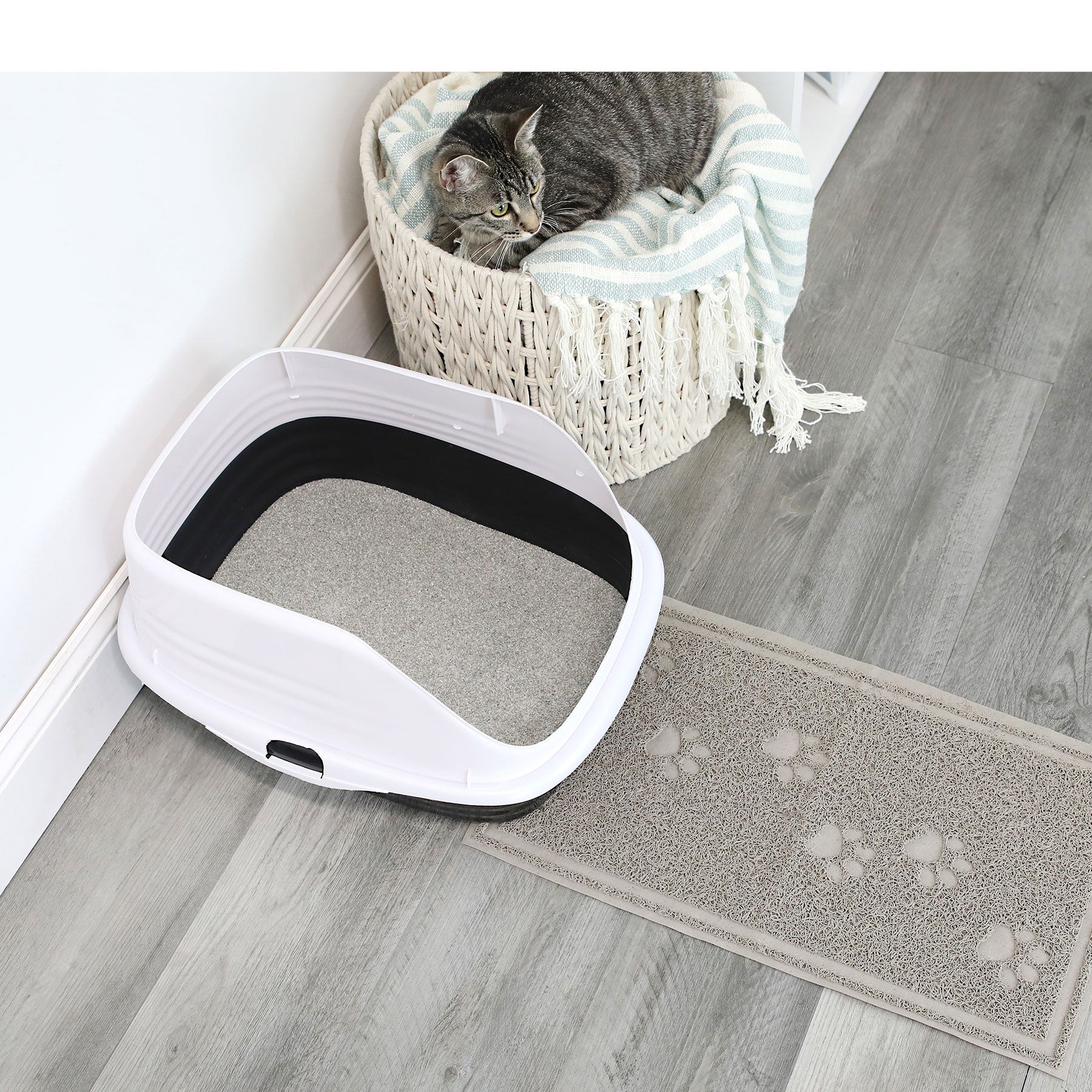 A gray tabby cat sits in a round basket with a striped blanket next to a white litter box filled with cat litter. The Arm & Hammer Paws Litter Mat is on the gray wooden floor, preventing scatter and showcasing its durable rubber material.