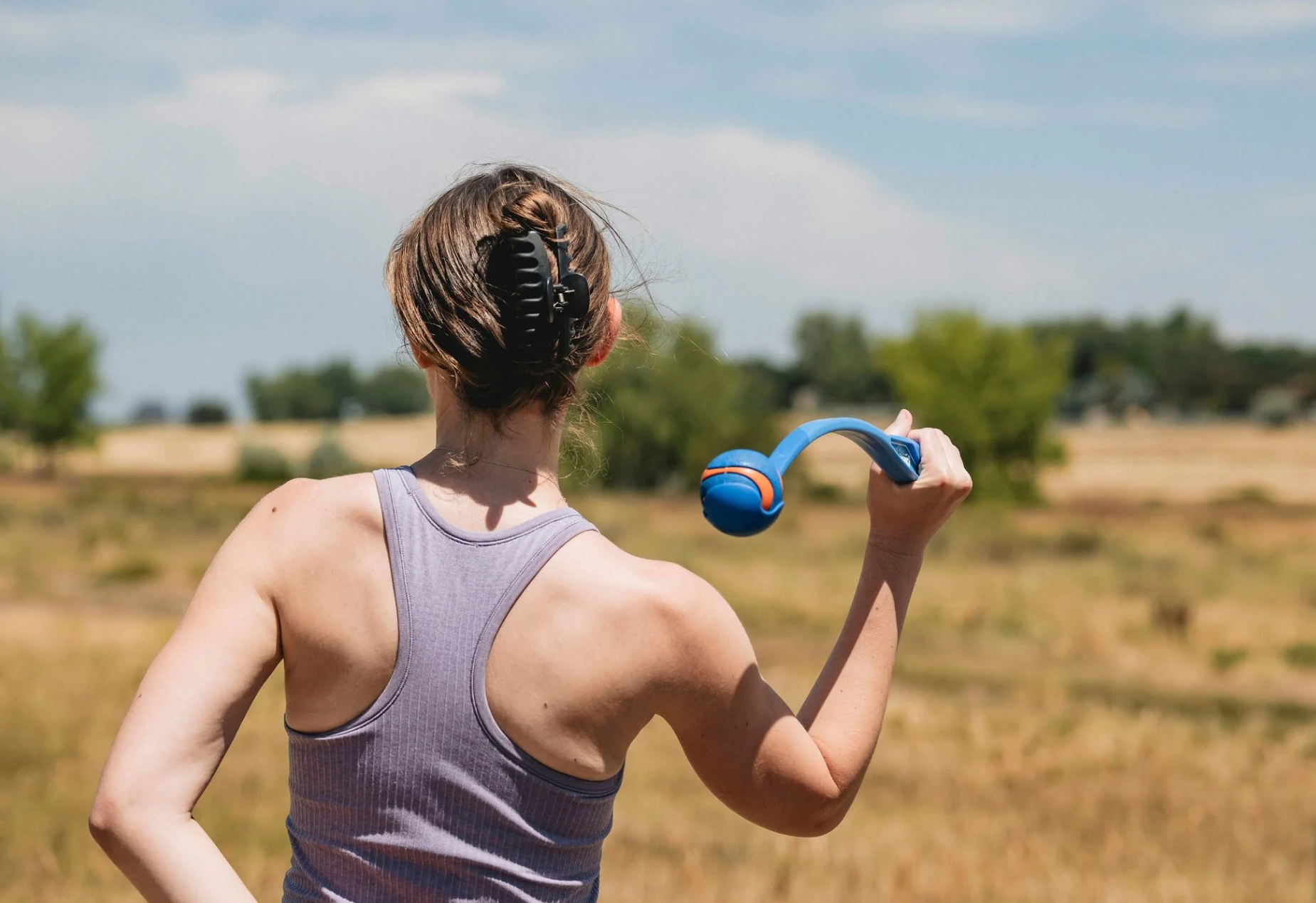 A woman is facing away from the camera, she is playing fetch outside with the Chuckit! Sport Launcher.