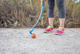 A person in gray leggings and pink shoes stands on a dirt path with the Chuckit! Classic Dog Ball Launcher & Fetch Medley Bundle, ready to throw fetch balls for a dog. Bushes and plants create the scenic backdrop for this playful moment.