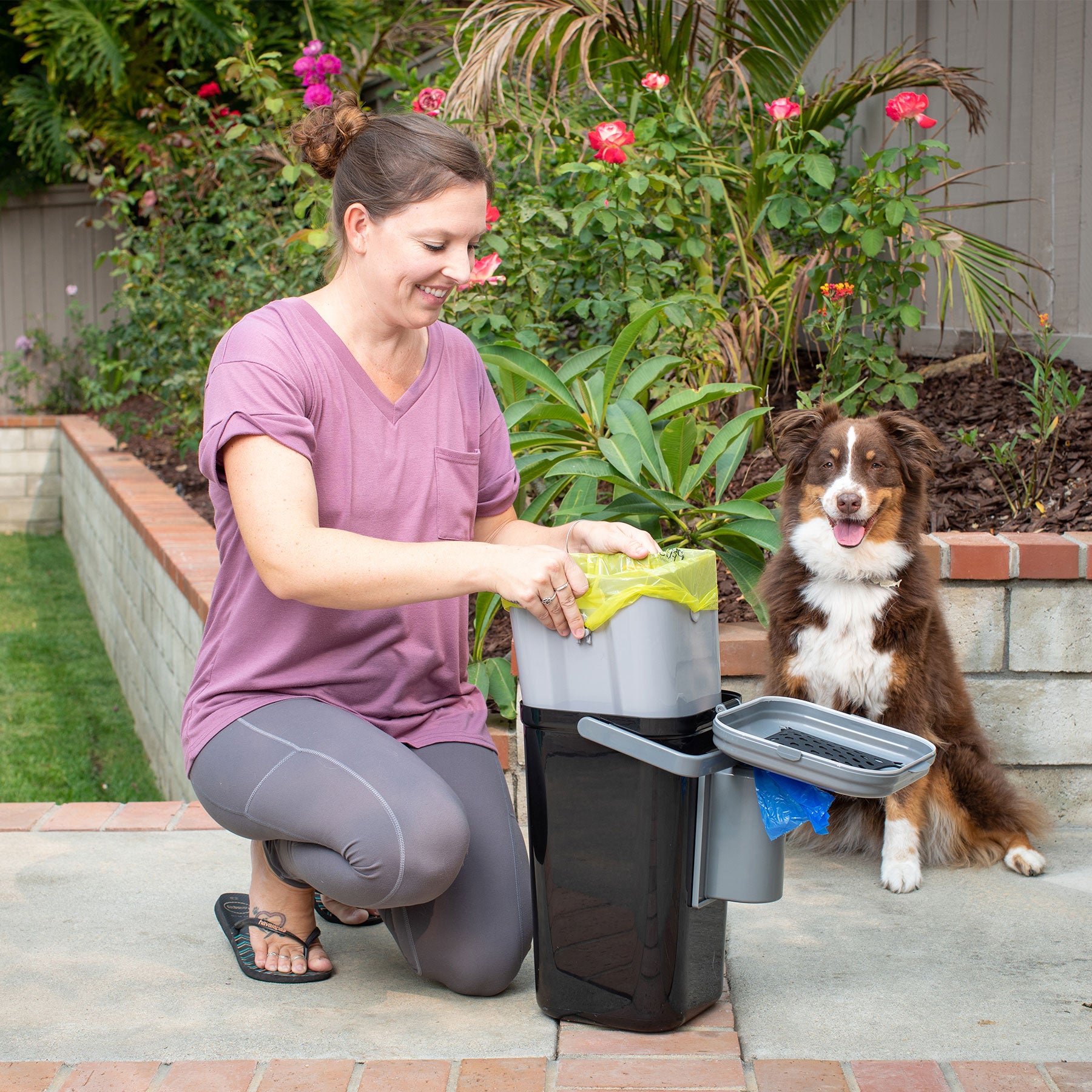 A woman kneels on a patio beside her panting dog, smiling as she uses the PetFusion Portable Outdoor Pet Waste Disposal System by Pet Fusion. Surrounded by colorful flowers and lush greenery, this pet waste station makes lawn clean-up easy.