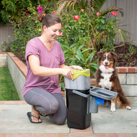 A woman kneels on a patio beside her panting dog, smiling as she uses the PetFusion Portable Outdoor Pet Waste Disposal System by Pet Fusion. Surrounded by colorful flowers and lush greenery, this pet waste station makes lawn clean-up easy.