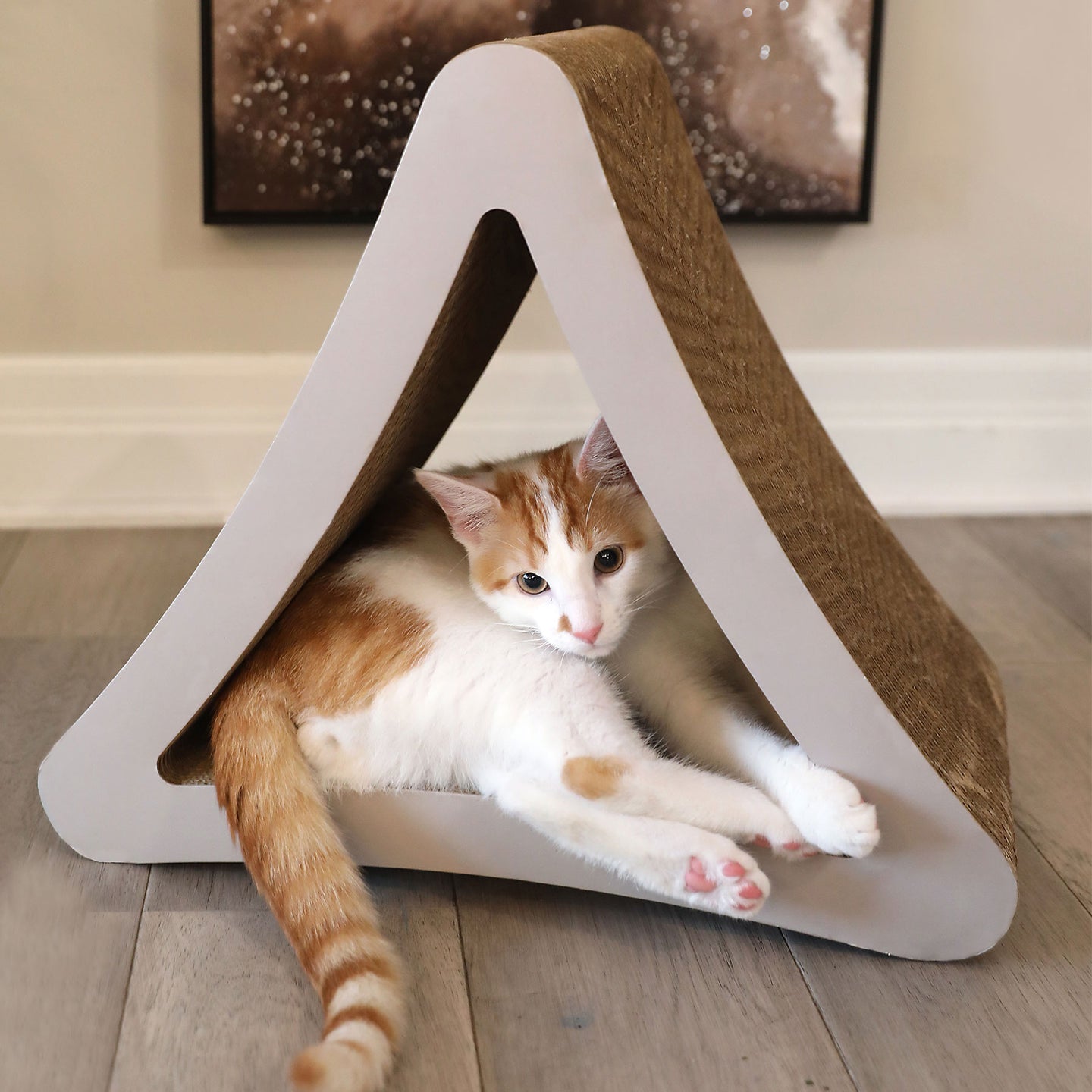 An orange and white cat relaxes in a Petfusion 3-Sided Vertical Cat Scratching Post, made of recycled cardboard. The eco-friendly post, set on a wooden floor, offers a scratchable textured surface with abstract art framed on the wall behind.