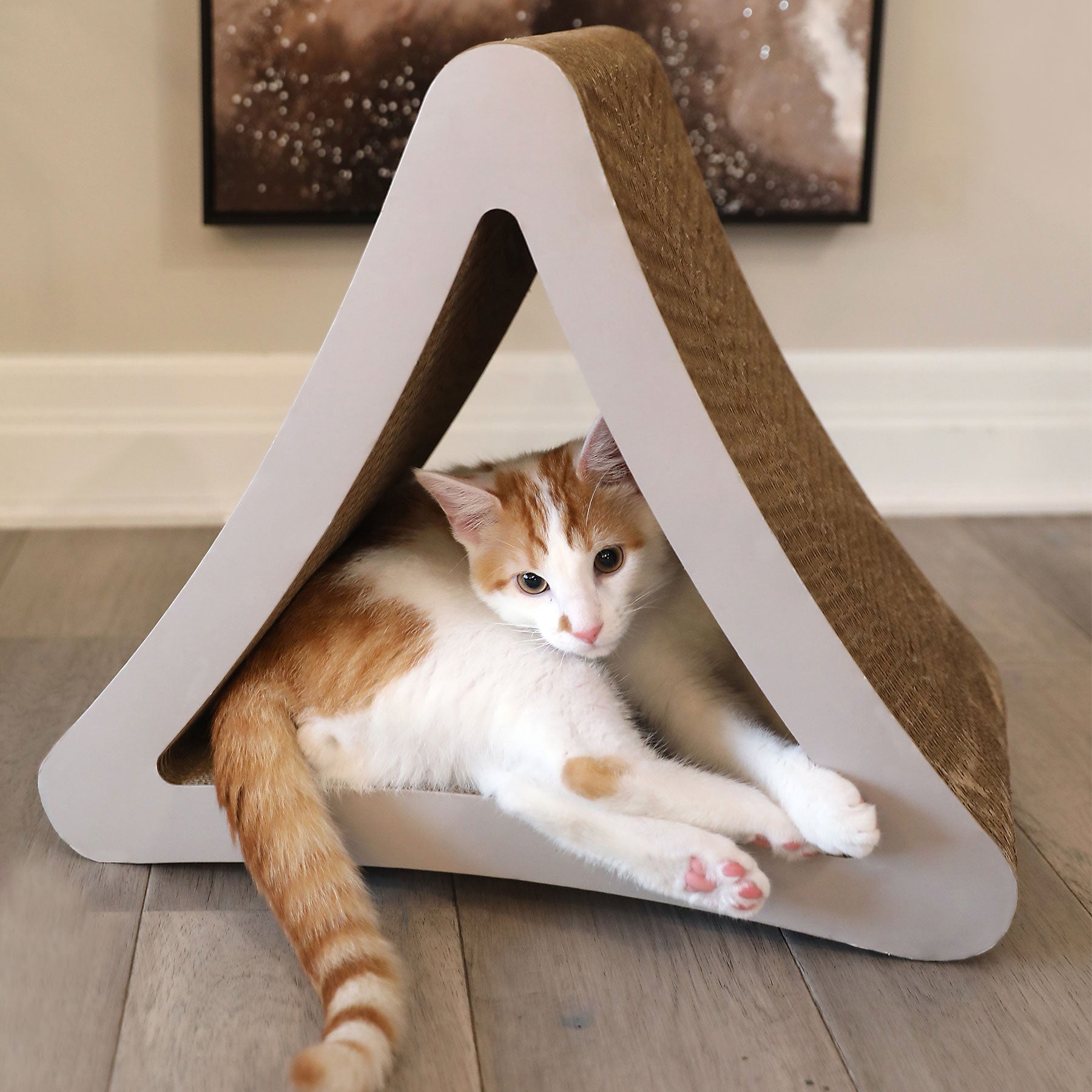 An orange and white cat relaxes in a Petfusion 3-Sided Vertical Cat Scratching Post, made of recycled cardboard. The eco-friendly post, set on a wooden floor, offers a scratchable textured surface with abstract art framed on the wall behind.