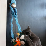 A gray cat engages with the Mad Cat Sushi Time Doorknob Toy, featuring a blue octopus attached to a handle. Its tentacles and colorful balls with feathers entice play, while bursts of silvervine heighten excitement.
