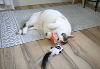 A fluffy white cat with brown patches playfully rolls on a gray patterned rug, clutching the Mad Cat Hiss & Haunt Plush Cat Toy in its paws. Nearby, a Halloween wand with a feathered tail enticingly lies on the wooden floor.