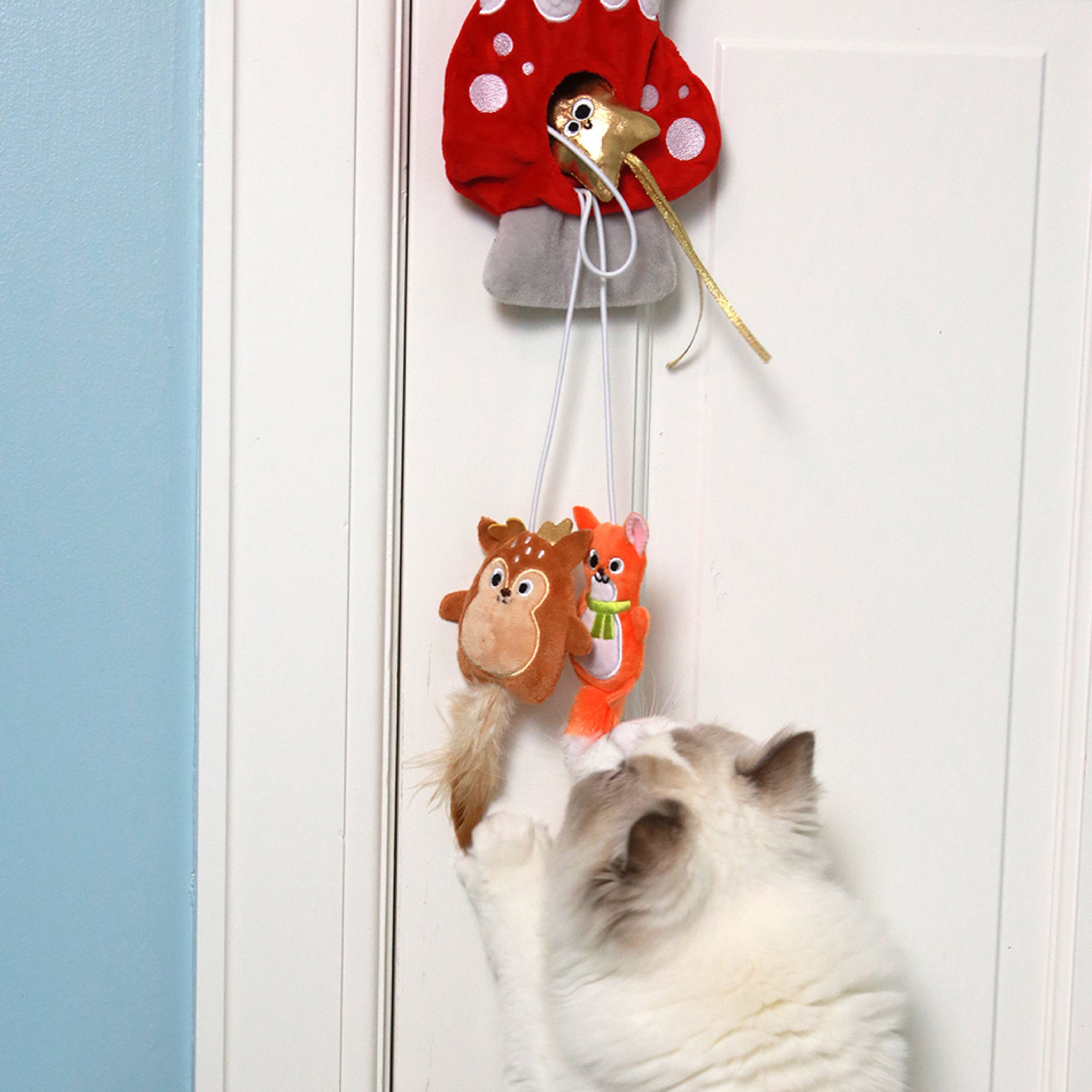 A cheerful white and gray cat stretches toward two plush toys—a brown squirrel and an orange fox—hanging from the Mad Cat Winter Woodland Doorknob Cat Toy, a playful red mushroom with white spots, part of a 3-pack designed to entertain any feline.