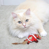 A fluffy cream-colored cat with blue eyes lounges on a carpet, playfully batting at a Mad Cat Ugly Sweater Stuffer Reindeer toy, complete with antlers and a feathered tail.
