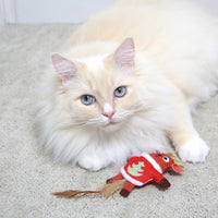 A fluffy cream-colored cat with blue eyes lounges on a carpet, playfully batting at a Mad Cat Ugly Sweater Stuffer Reindeer toy, complete with antlers and a feathered tail.