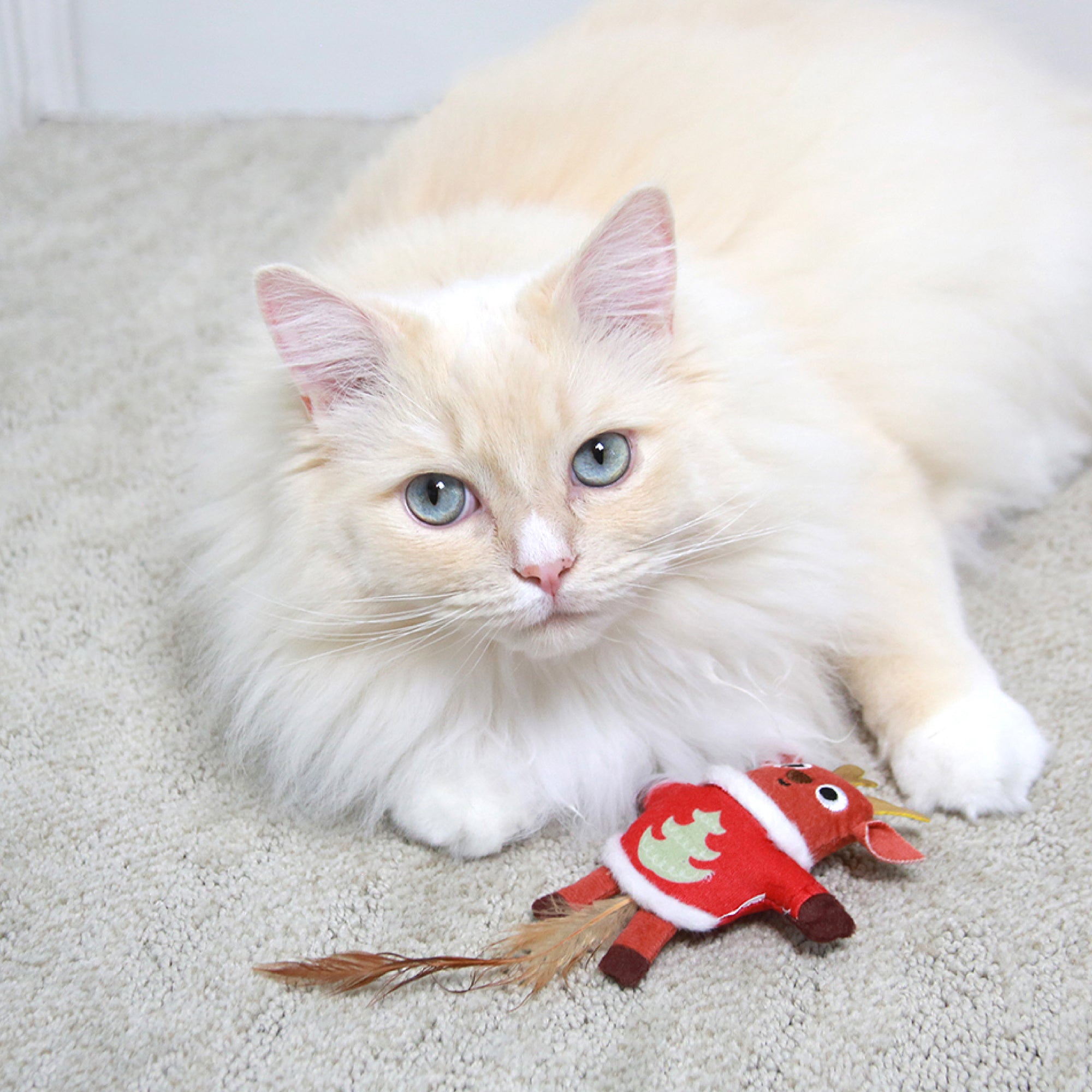 A fluffy cream-colored cat with blue eyes lounges on a carpet, playfully batting at a Mad Cat Ugly Sweater Stuffer Reindeer toy, complete with antlers and a feathered tail.