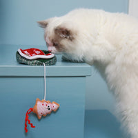 A fluffy white cat, reminiscent of the Mad Cat Holiday Cookie Tin Hide A Toy, curiously sniffs a smiling fish-shaped fabric toy attached by a string to a small red cushion against a light blue background.