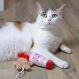 A white and orange calico cat with green eyes lies on a wooden floor, exuding festive charm like Mad Cat Santa Claws Milk. One paw rests on a bottle-shaped toy, while a plush gingerbread man is beside it. The cat gazes up expectantly, as if waiting for milk to magically appear.