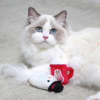 A fluffy white and gray cat with blue eyes relaxes on a light carpet surrounded by plush toys. Among them are Mad Cats Cool Cocoa Cat Toy, featuring a snowman and red mug with gingerbread man designs, perfect for cute mad cat moments.