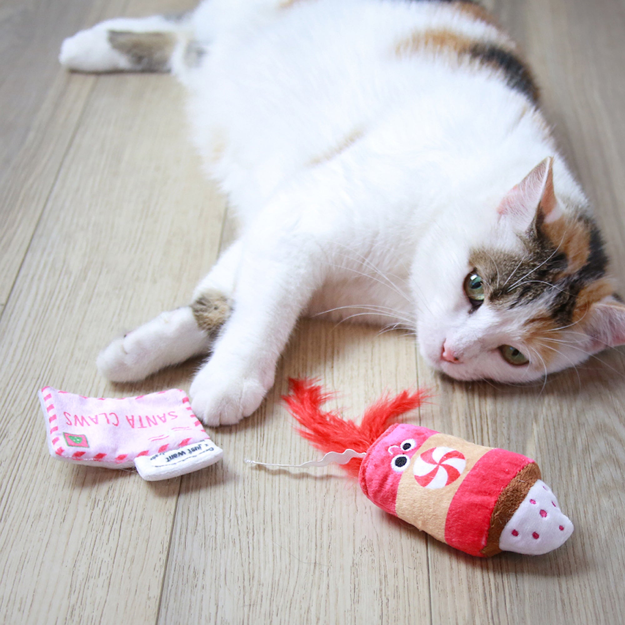 A white and brown cat lounges on a wooden floor next to two Mad Cat toys: one shaped like a mug with Santa Claws, nodding to a Pepurrmint Latte, and the other resembling a peppermint-themed treat with a red feather.