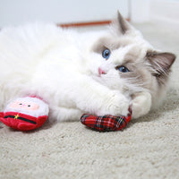 A fluffy white and gray cat with blue eyes relaxes on a carpet, cuddling a red plaid heart-shaped toy and a Santa-themed plush from the Mad Cat Oh So Jolly 3 Pack.