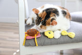 A calico cat lounges on a gray cushioned chair, resting among three Quirky Kitty Pasta Pals catnip toys—a brown one with checkered legs, a yellow chick-shaped toy, and one with a yellow bow—from Quirky Kitty.