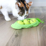 A white and brown cat sniffs the Quirky Kitty Pea Pawd Kicker – a large catnip kicker toy by Quirky Kitty, shaped like a pea pod with two smiling peas inside, lying on a wooden floor.