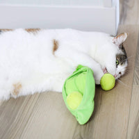 A white cat with brown spots lies on a wooden floor, playfully biting the Quirky Kitty Pea Pawd Kicker – a large catnip kicker toy shaped like a pea pod with two tennis ball-like peas, by Quirky Kitty.