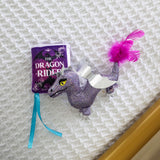 The Quirky Kitty Fairy Tail Romance 2pk Cat Toy Set with Catnip features a plush dragon toy with silver wings and a purple feathered tail next to a purple fabric book, perfect for fairy tale-loving cats.