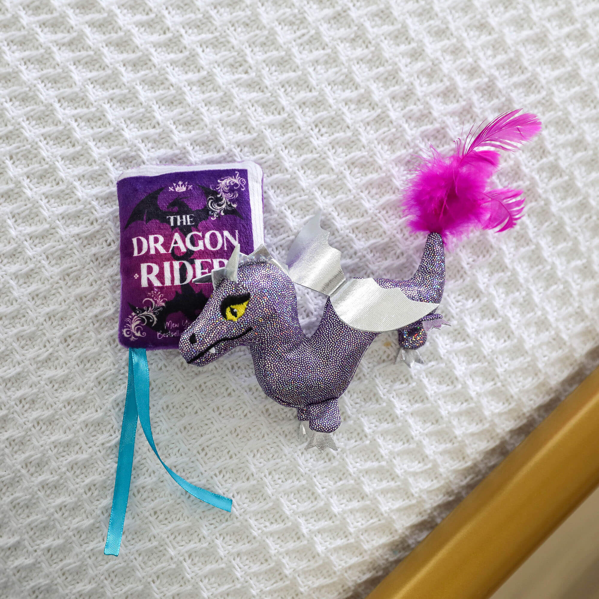 The Quirky Kitty Fairy Tail Romance 2pk Cat Toy Set with Catnip features a plush dragon toy with silver wings and a purple feathered tail next to a purple fabric book, perfect for fairy tale-loving cats.