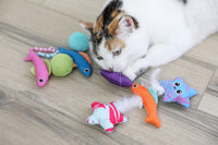 A white and orange cat lies on a wooden floor, playing with the Quirky Kitty Fish Bowl 8pk—ocean-themed cooling cat toys with catnip from Quirky Kitty, featuring colorful fish, balls, and star shapes.