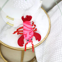 The Quirky Kitty Fresh Lobster Kicker—a catnip-filled, ocean-themed toy—rests on a marble-topped table with gold trim, beside a white textured blanket and stacked white boxes.