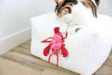 A calico cat sits on a white textured ottoman, playing with the Quirky Kitty Fresh Lobster Kicker—an ocean-themed, catnip-packed cat toy from Quirky Kitty—that dangles over the edge onto a wooden floor.
