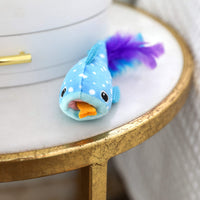 The Quirky Kitty Whale Shark Chomper – Cooling Ocean Cat Toy with Catnip rests on a gold-edged table next to a white object.