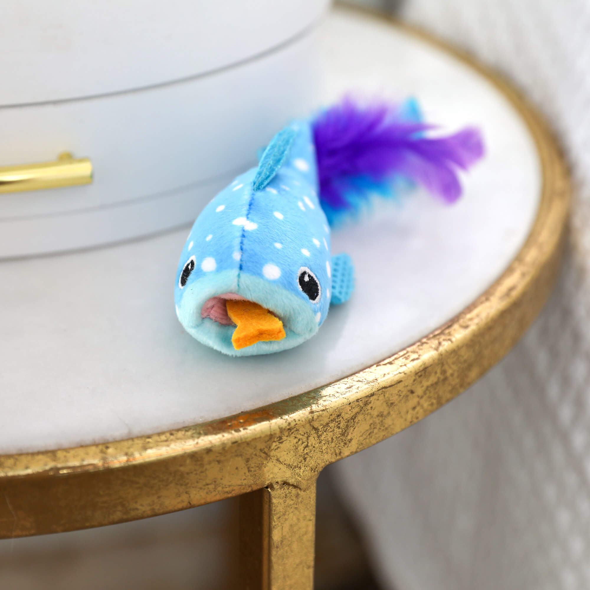 The Quirky Kitty Whale Shark Chomper – Cooling Ocean Cat Toy with Catnip rests on a gold-edged table next to a white object.