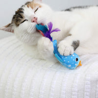 A white and tabby cat lies on a textured white blanket, playfully biting and holding the Quirky Kitty Whale Shark Chomper – a cooling ocean cat toy with catnip from Quirky Kitty, featuring fun purple feathers.