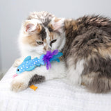 A fluffy calico cat lounges on a white textured surface, resting its head on the Quirky Kitty Whale Shark Chomper – Cooling Ocean Cat Toy with Catnip, gazing slightly to the side.