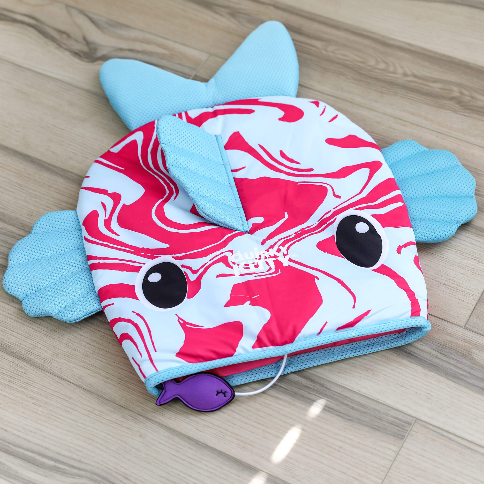 The Quirky Kitty Fish Cooling Mat – Ocean-Themed Cat Play Mat by Quirky Kitty features a red and white swirled fish design, blue fins, cartoon eyes, and a small attached purple toy—perfect for keeping cats cool on any wooden floor.