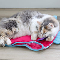 A fluffy gray and white cat relaxes on a wooden floor, lying on its side with its paw and head resting on the Quirky Kitty Fish Cooling Mat – an ocean-themed cat play mat by Quirky Kitty.