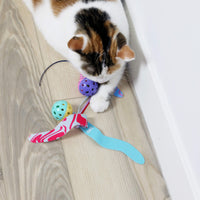 A white and brown cat plays on a light wooden floor with Quirky Kitty Catnip Seaweed Tumblers 2pk—ocean-themed cat toys with catnip—and colorful balls, next to a white wall.