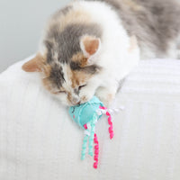 A fluffy cat lies on a white blanket, resting its face on the Quirky Kitty Jellyfish & Chill 2-Pack Cat Toy with Catnip by Quirky Kitty, featuring blue and pink jellyfish details with ribbons.