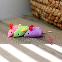 Three Quirky Kitty Glow Mice—catnip & silvervine-filled cat toys in pink, green, and purple—sit on the floor near a basket as sunlight streams in, perfect for interactive nighttime play.