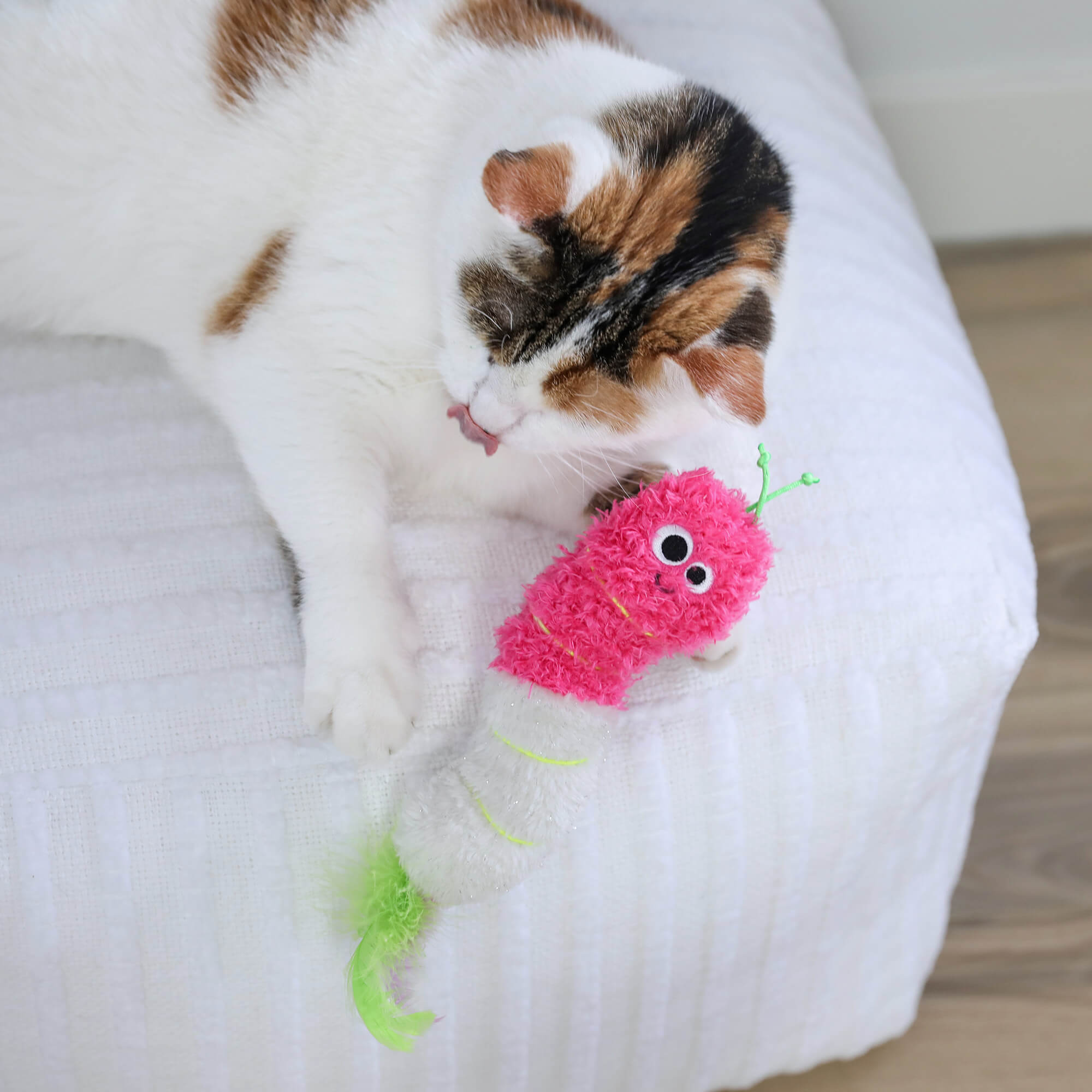 A white, brown, and black cat is lying on a white textured surface licking its paw next to the Quirky Kitty Glow Worm Kicker—a pink and white catnip and silvervine toy by Quirky Kitty that glows at night with playful details.