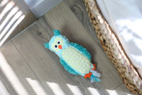 The Quirky Kitty Night Owl Kicker – a turquoise and yellow glow-in-the-dark catnip & silvervine toy shaped like an owl – rests on a light wooden floor near a woven basket and bed, with sunlight casting striped shadows.